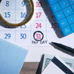 Calendar page marking payday on the 31st with a circled date, a blue calculator, a magnifying glass, a black pen, and cash nearby.