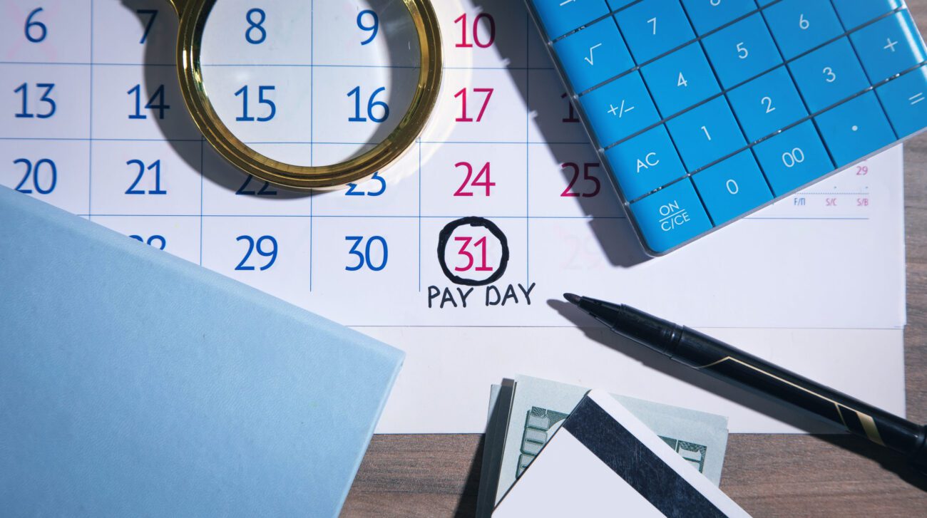 Calendar page marking payday on the 31st with a circled date, a blue calculator, a magnifying glass, a black pen, and cash nearby.