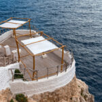 Clifftop terrace with whitewashed walls and wooden railings, shaded by white fabric canopies, overlooking the blue ocean.