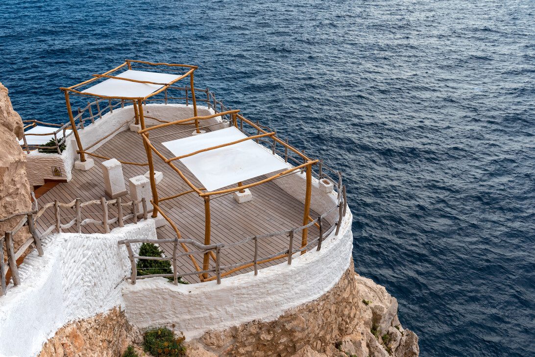 775289 Expomobles | diario-economia.com Clifftop terrace with whitewashed walls and wooden railings, shaded by white fabric canopies, overlooking the blue ocean.