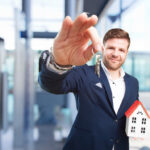 Businessman in a navy suit extends key ring toward the camera, holding a small white house model with a red roof as a symbol of home ownership and real estate.