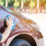 Person wiping a maroon car’s hood with a blue microfiber cloth during a car wash or detailing session
