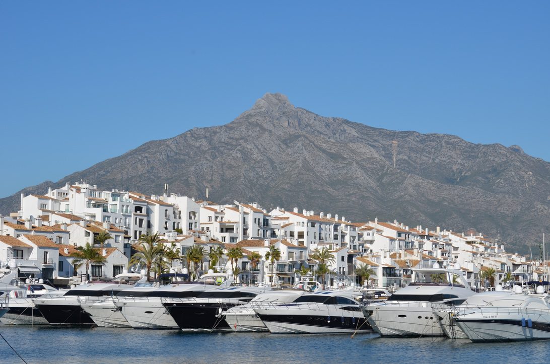775802 - Absolute Prestige 1 | diario-economia.com White coastal town with terracotta rooftops and palm trees beside a marina full of luxury yachts, mountain backdrop.
