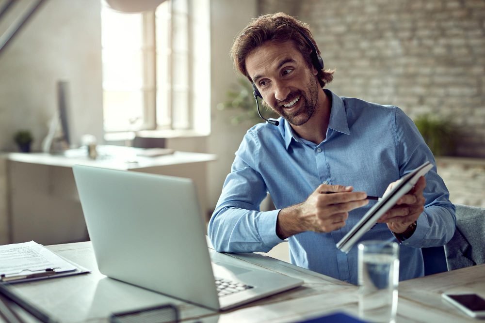 776006 - Aselai Asesores | diario-economia.com Man wearing a headset at a desk with a laptop and tablet, smiling during a call or presentation in a bright office.