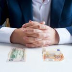 Businessperson with clasped hands on a table; a US $50 bill and a €50 note lie in front, suggesting a payment or bribe.