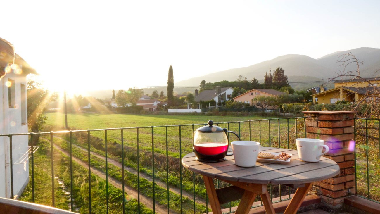 Sunlit balcony table at sunrise with a glass teapot of red tea, two white cups, and a plate of cookies over a green field and distant houses.
