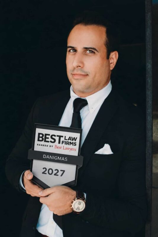 danigmas - IP | diario-economia.com Professional man in a black suit and tie holding a 'Best Law Firms' award plaque that reads DANIGMAS, 2027, against a dark background.