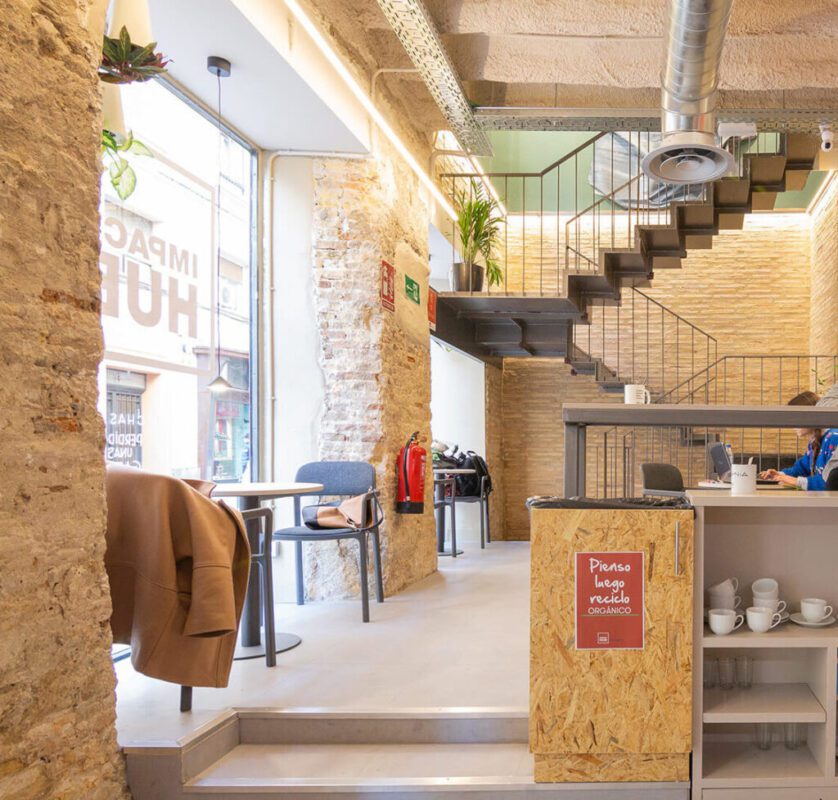 775783 - HUS Proyectos | diario-economia.com Bright industrial café interior with exposed brick, a metal staircase, and a service counter with cups.