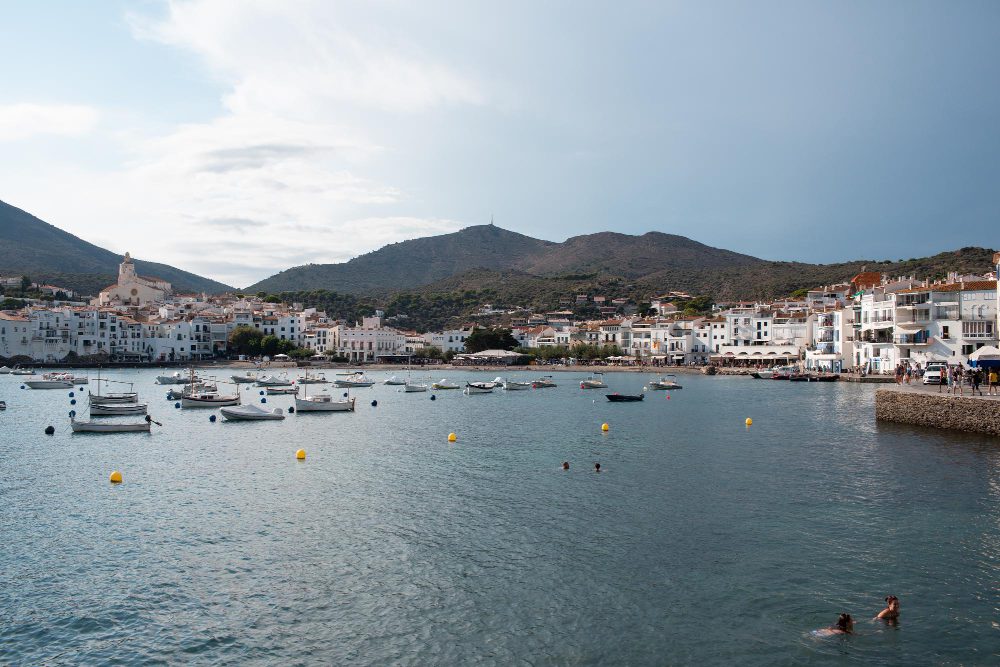White-washed coastal town along a calm harbor with moored boats and hills in the background.