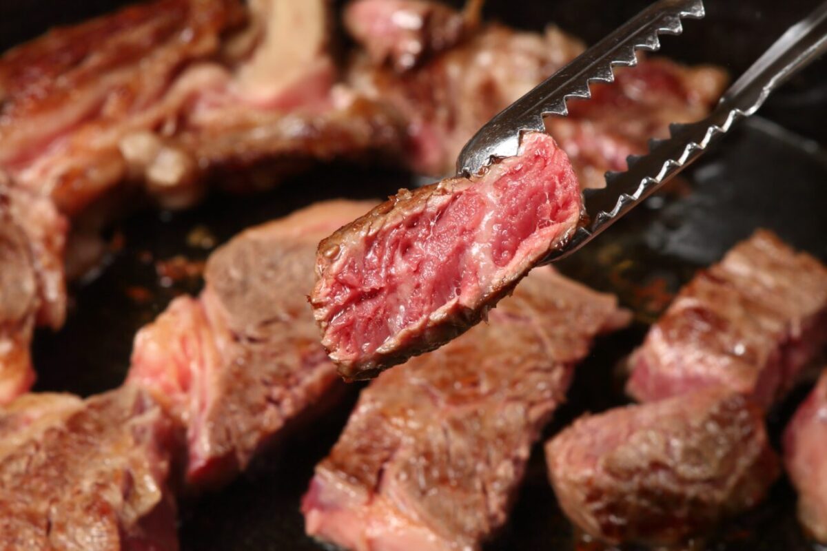 Medium-rare steak pieces in a skillet with a pair of tongs lifting a pink center slice into view