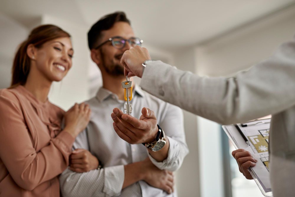 776606 - Gestibensa | diario-economia.com Smiling couple receives house keys from a real estate agent in a bright office setting.