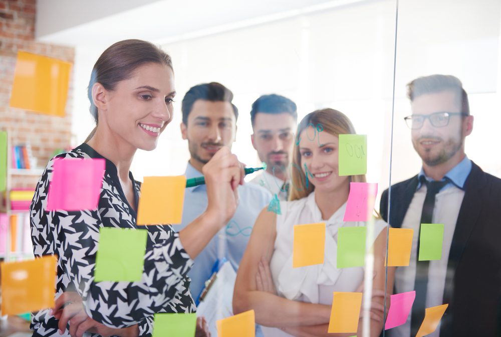 776635 - Talent Swarm | diario-economia.com Team of five coworkers collaborating at a glass wall covered in colorful sticky notes during a brainstorming session.