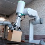 Industrial robot arm lifting a cardboard box off a conveyor belt in a tech facility with grey brick walls and overhead lighting.