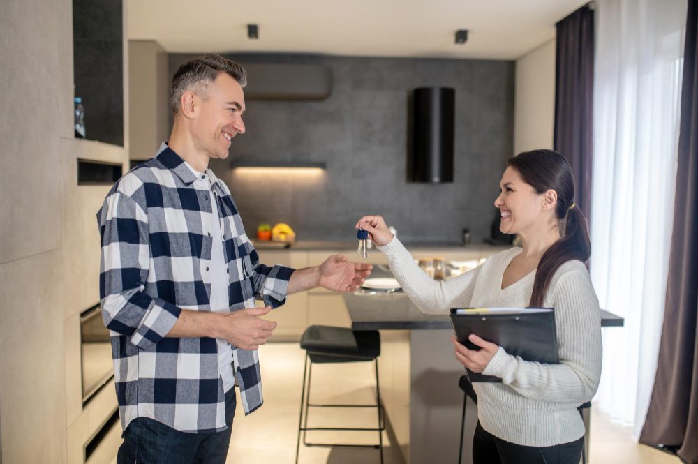 Real estate agent hands keys to a smiling man in a modern kitchen setting during a home handover.