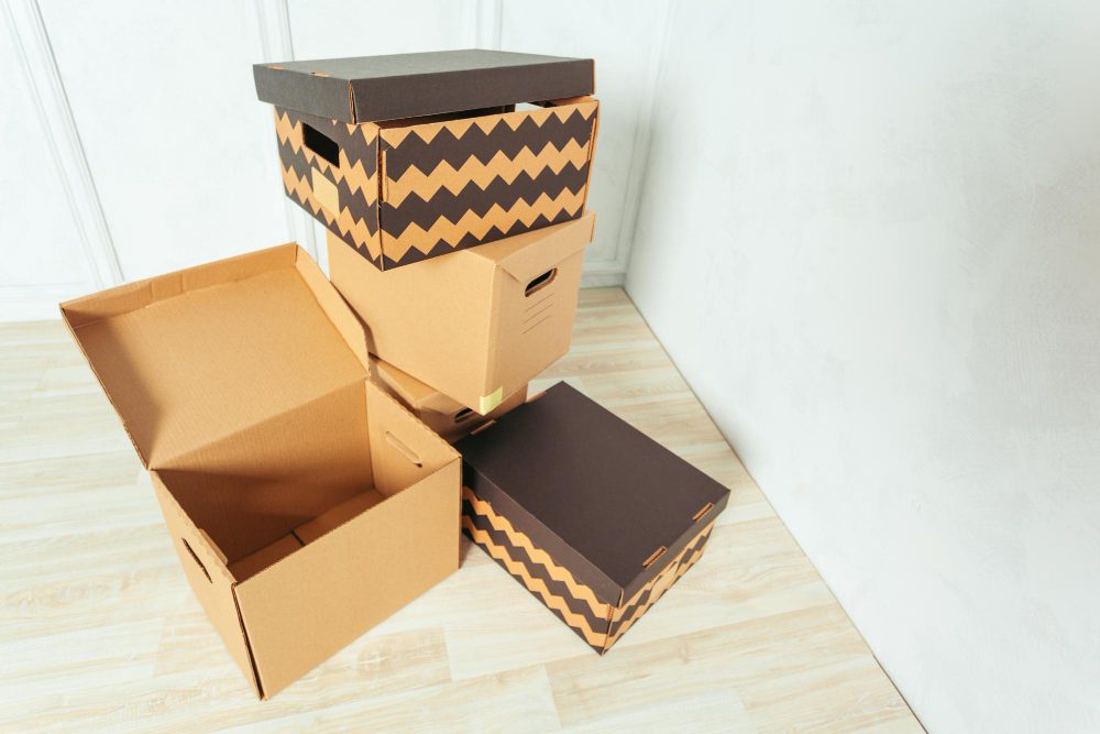 777899 - Cartonajes VAHL | diario-economia.com Stack of cardboard moving boxes in a corner, some open and others closed, in a minimal room with wooden floor.
