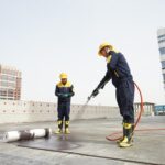 Two construction workers in yellow helmets and navy uniforms on a rooftop; one uses a hose while the other watches nearby.