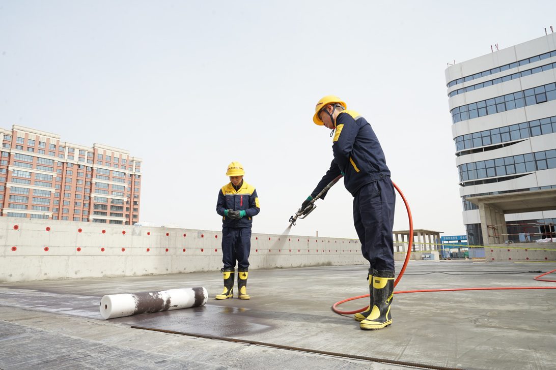 777977 - CINAT | diario-economia.com Two construction workers in yellow helmets and navy uniforms on a rooftop; one uses a hose while the other watches nearby.