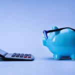 Blue piggy bank wearing glasses beside a handheld calculator on a blue surface, symbolizing savings and budgeting.