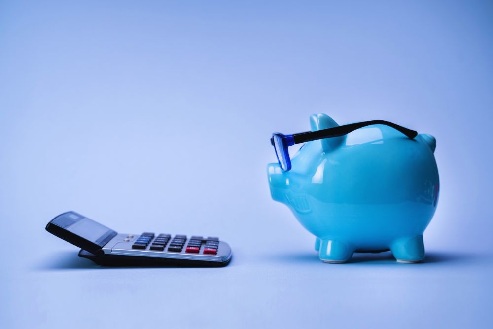 777984 - Consulae | diario-economia.com Blue piggy bank wearing glasses beside a handheld calculator on a blue surface, symbolizing savings and budgeting.
