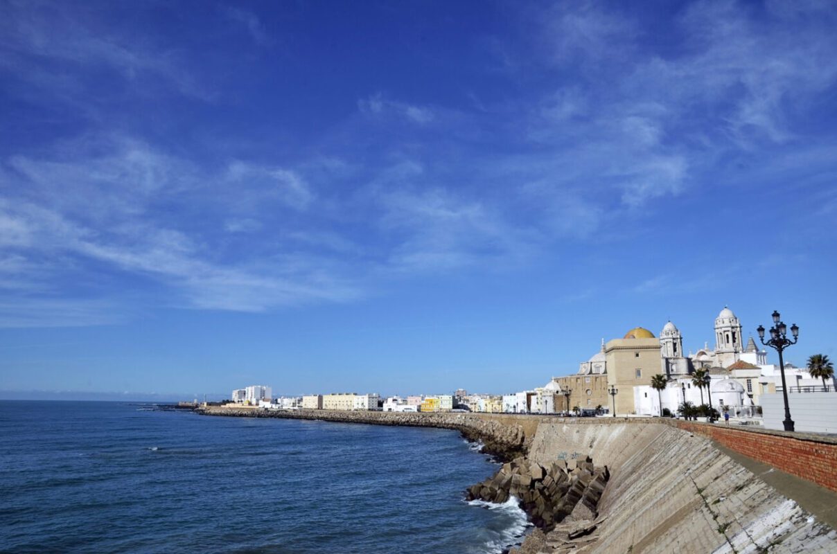 CADIZ SPAIN | diario-economia.com Coastal town along a breakwater with white and beige buildings and a domed church against a blue sky.