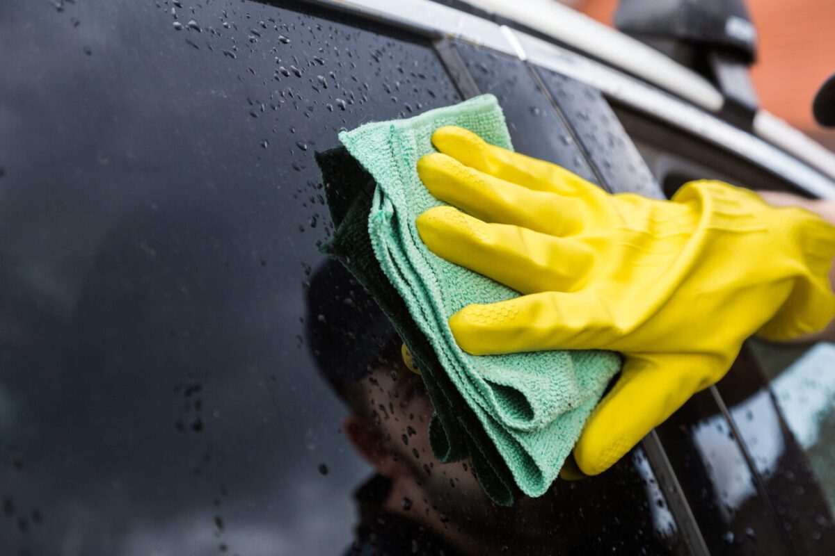 Person wearing yellow gloves wiping a car window with a green microfiber cloth, water droplets on the glass.
