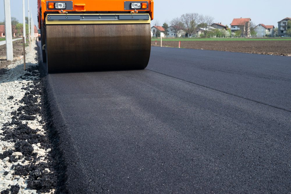 778216 - INTRAME | diario-economia.com Orange road roller compacting a fresh layer of asphalt on a newly paved road, with a gravel shoulder on the left and houses in the background.