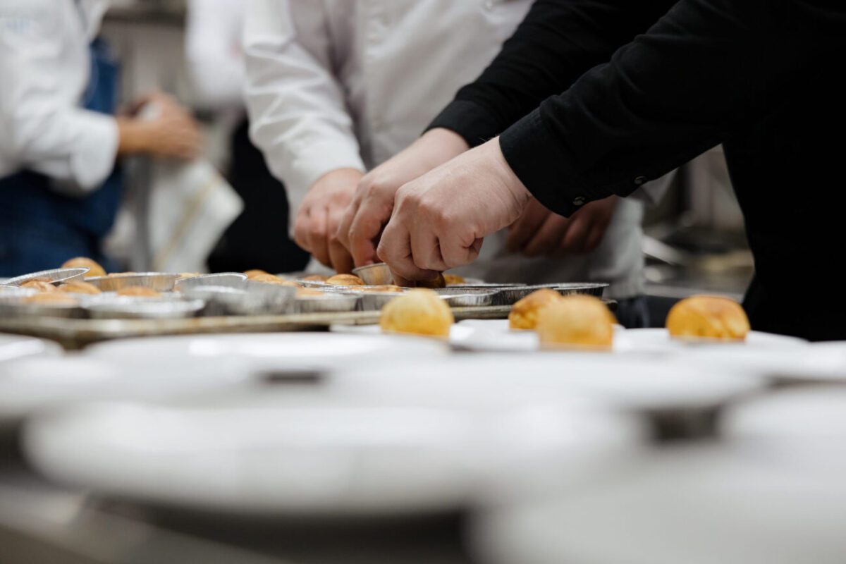 Hands of several chefs arranging small pastries on metal trays in a busy kitchen setting in white coats and black sleeves, focusing on the prep line.