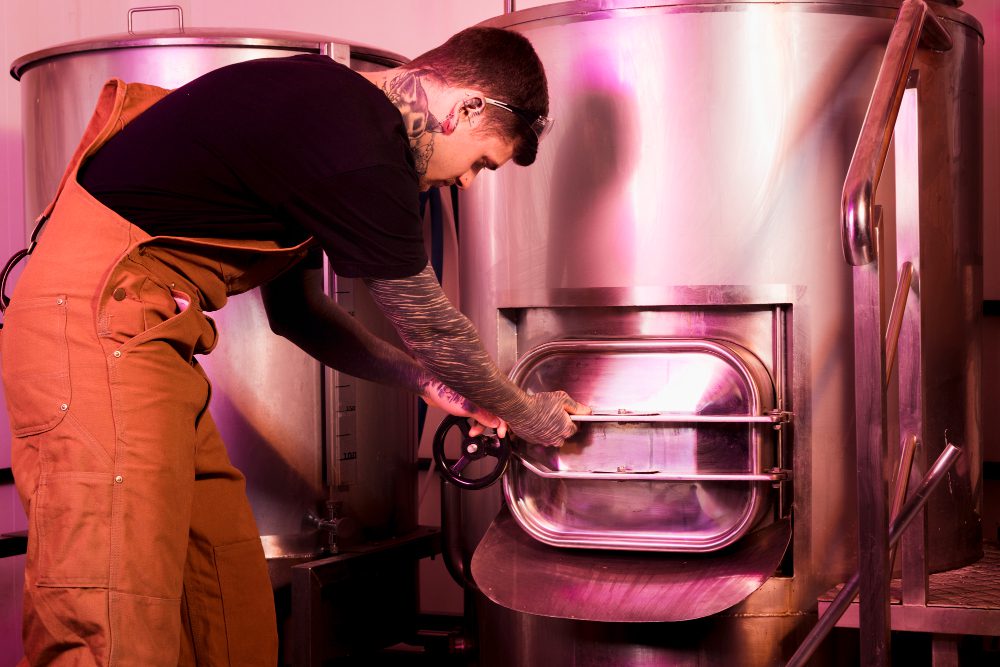778477 - Multifiltra | diario-economia.com Man in brown overalls opens a large stainless-steel industrial kettle, turning the front valve by its handwheel.