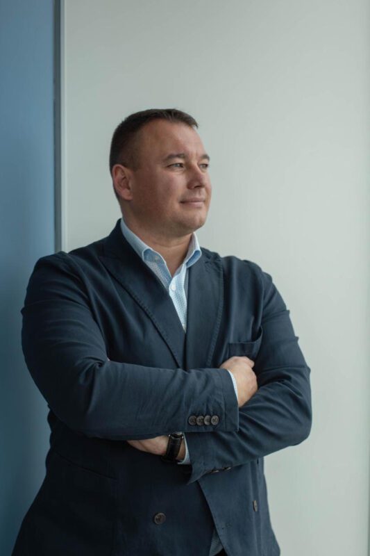 Man in a dark blazer standing with arms crossed, looking pensively to the right by a light wall.
