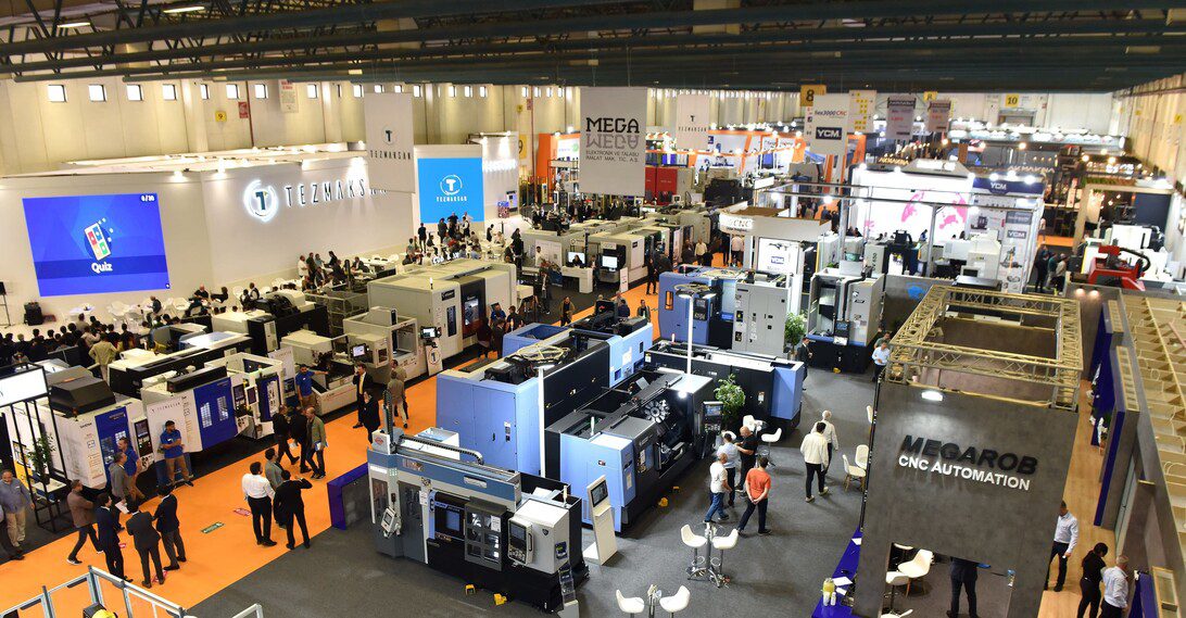 A large industrial trade show hall filled with CNC machines and exhibitor booths, attendees mingle among blue and gray machinery displays.