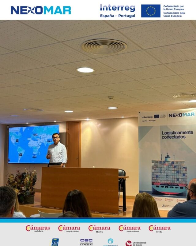 Presenter at a podium giving a conference talk on logistics, with a world map on a screen behind him and an attentive audience.