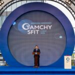 Speaker at a podium on a blue circular-stage backdrop for the Opening Ceremony (text on screen reads 'Opening Ceremony GAMCHY SFIT 3 July 2026')