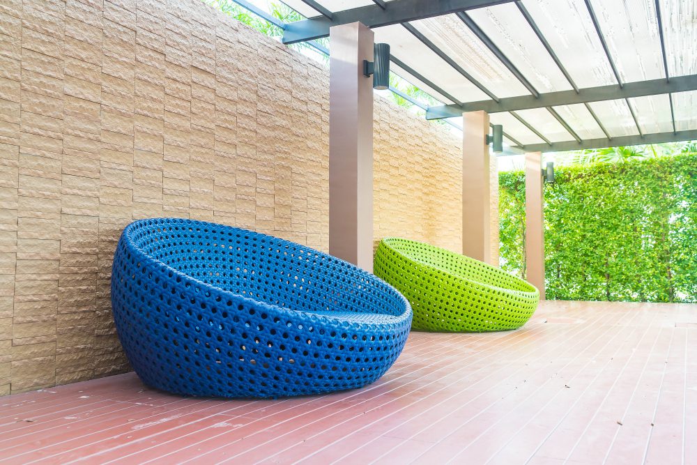 778533 - Toldos Eguzki | diario-economia.com Blue and green woven circular lounge chairs on a wooden deck under a glass-roofed patio with a brick wall and green hedge in the background.