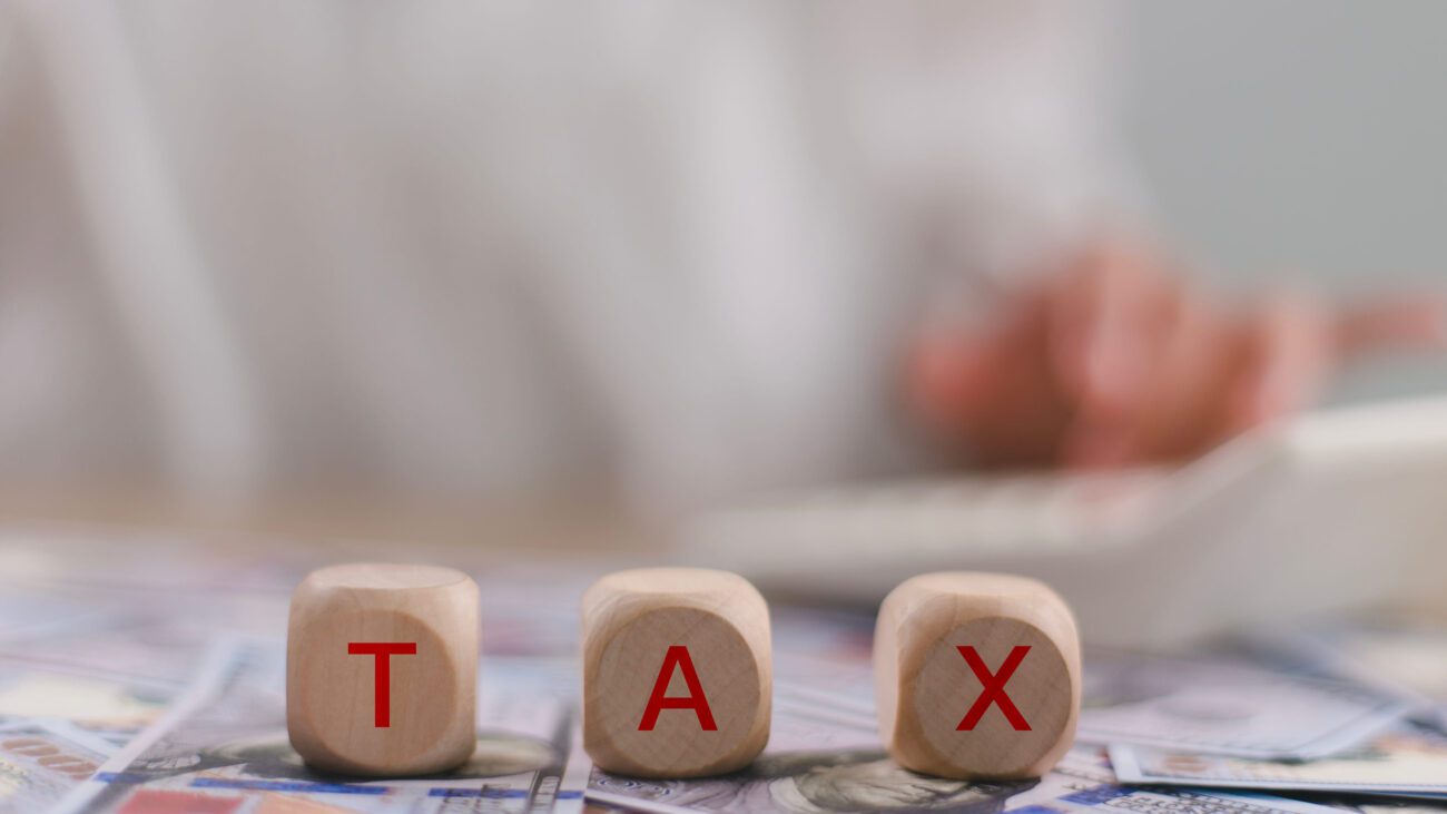 Tax word on wooden cubes with a woman working in the background | diario-economia.com Tax concept: three wooden blocks spell TAX atop scattered cash, foreground in focus, background blurred.