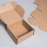 Open brown cardboard box with its lid raised, and flat corrugated panels lying nearby on a gray surface.