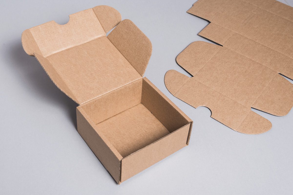 779277 - Letro | diario-economia.com Open brown cardboard box with its lid raised, and flat corrugated panels lying nearby on a gray surface.