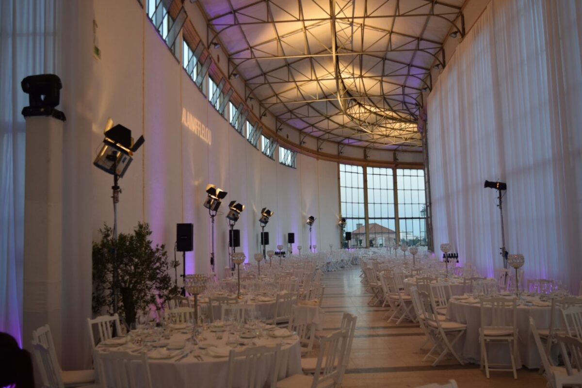 Banquet hall set for a formal event with round tables, white chairs, and purple uplighting along tall white curtains.