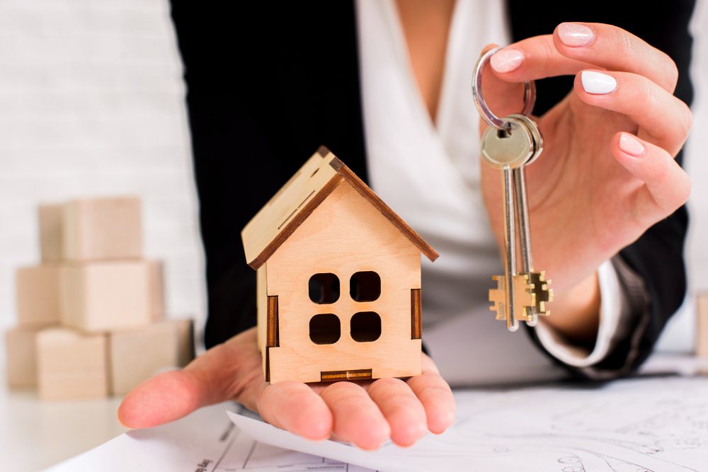 780370 - Finques Sant Pol | diario-economia.com Person in business attire holds a small wooden house model and a key, symbolizing home ownership or real estate