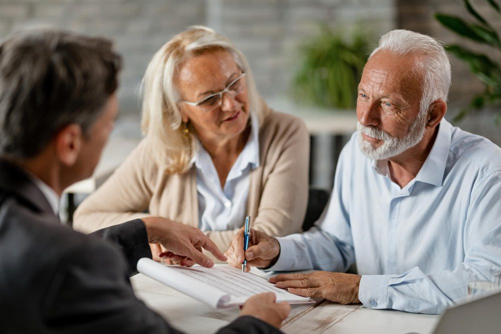 780500 - Devengo Abogados | diario-economia.com Two older adults sign a document with a man in a suit guiding them at a bright table in a professional setting.