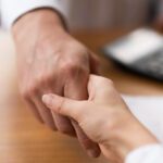 Two people shake hands across a wooden desk, signaling agreement or a handshake in a business meeting.