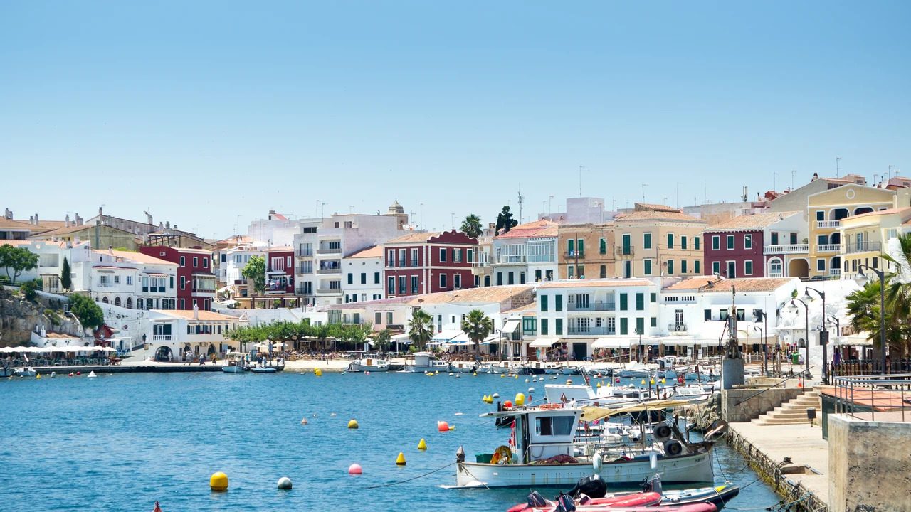 757555 - Menorca Secret | diario-economia.com Harbor scene in a Mediterranean town with colorful buildings along the waterfront and boats anchored in calm blue water