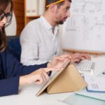 Two colleagues in a bright office; the woman taps a tablet resting on a beige stand while wearing glasses and earbuds as the man works on a laptop nearby.