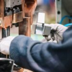 Worker in gloves operating a metal stamping machine, guiding a silver block into place between copper-colored components