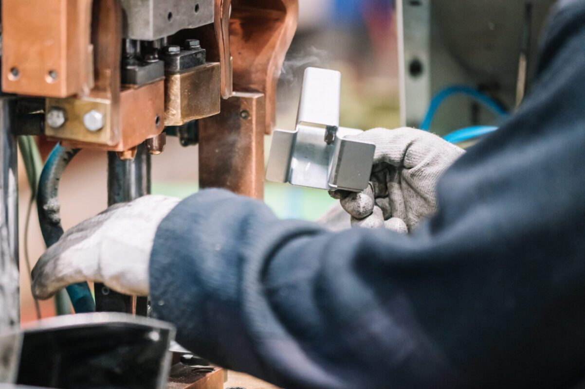 780423 Metric 4 | diario-economia.com Worker in gloves operating a metal stamping machine, guiding a silver block into place between copper-colored components