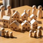 A circle of wooden figurines surrounds several small wooden houses on a table, depicting community housing or neighborhood unity.