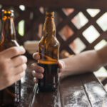 Hands hold brown beer bottles around a glossy wooden table on a sunny deck with lattice in the background, suggesting a casual outdoor gathering.