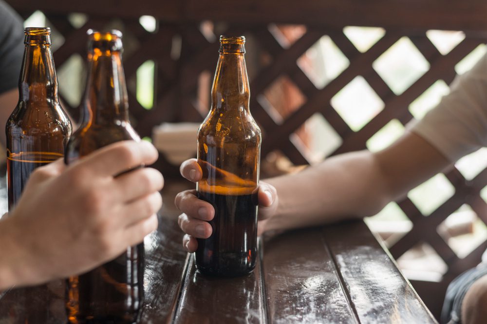 Hands hold brown beer bottles around a glossy wooden table on a sunny deck with lattice in the background, suggesting a casual outdoor gathering.