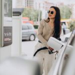 Woman in a beige cardigan and sunglasses refueling a car at a gas station.