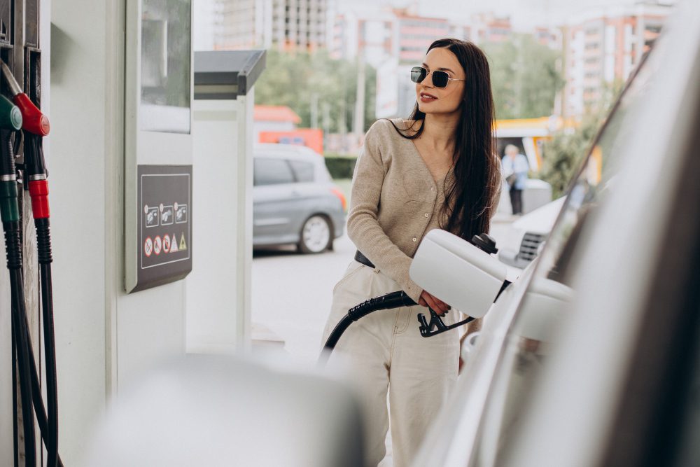 Woman in a beige cardigan and sunglasses refueling a car at a gas station.