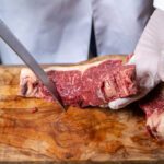 Butcher wearing gloves slicing a raw beef steak on a wooden cutting board with a large knife